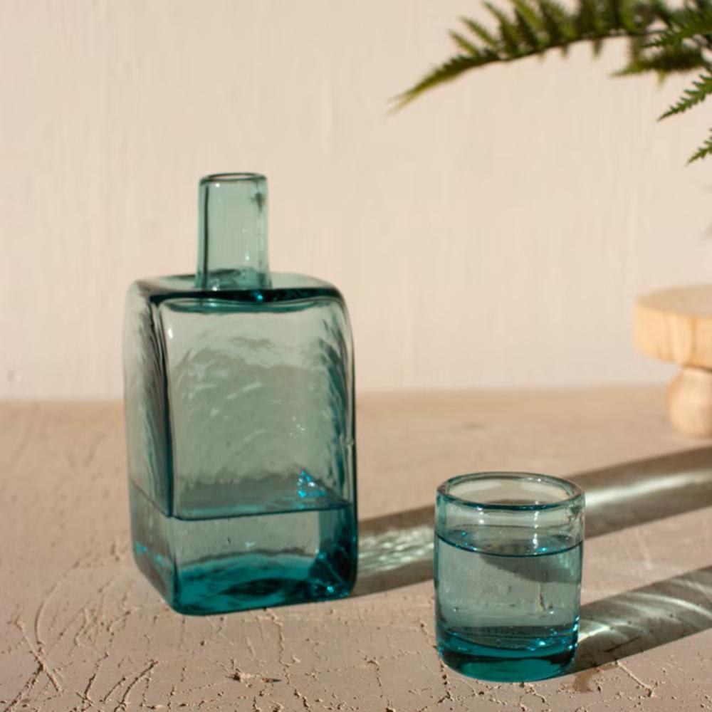 A translucent teal blue glass decanter with a matching small tumbler, styled on a textured beige stone surface with warm sunlight and shadows.