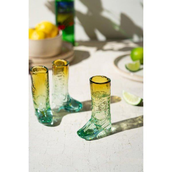 Three green glass boot-shaped shot glasses on a light surface with lemons and limes in the background.
