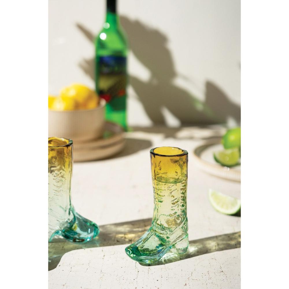 Two glass boot-shaped tumblers on a table with lemons and a bottle in the background.