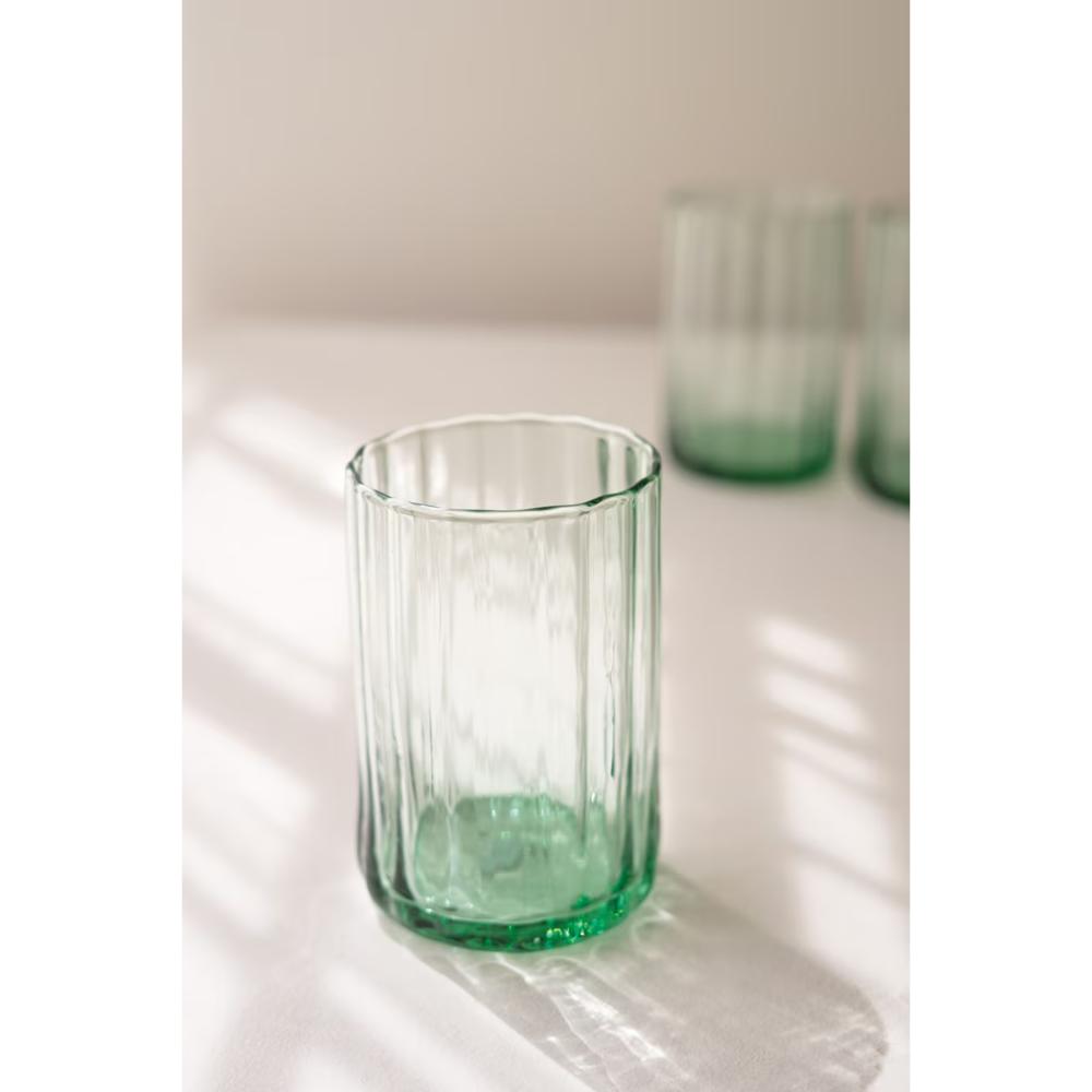A clear glass tumbler with a green tint and vertical fluted texture, sitting on a white surface with soft window shadows. Two more matching glasses are blurred in the background.