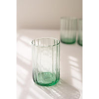 A clear glass tumbler with a green tint and vertical fluted texture, sitting on a white surface with soft window shadows. Two more matching glasses are blurred in the background.