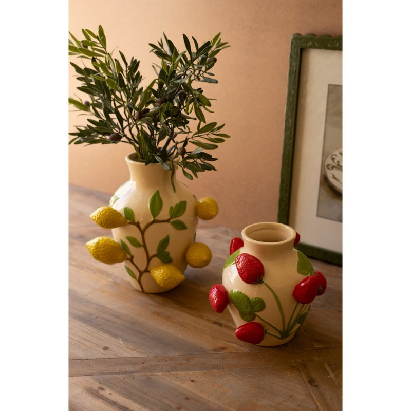 Two Kalalou vases on a wooden table, one with yellow fruit, the other red fruit designs, filled with greenery.