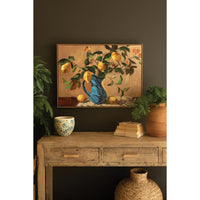 A framed painting of lemons in a blue vase hangs on a dark brown wall above a rustic wooden console table styled with plants, a ceramic bowl, and woven decor.