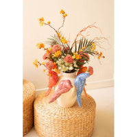 Decorative vase with birds and colorful flowers on a woven stool against a plain wall.