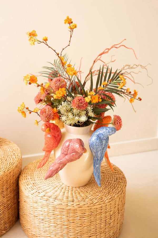 Decorative vase with birds and colorful flowers on a woven stool against a plain wall.