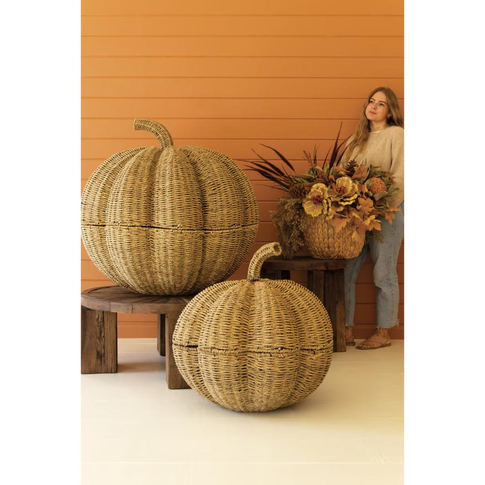Two large woven pumpkin-shaped decorative containers with lids are displayed on wooden stools in front of an orange wood-paneled wall. A woman stands behind holding a floral arrangement, with one pumpkin container in the foreground and another larger one behind it.
