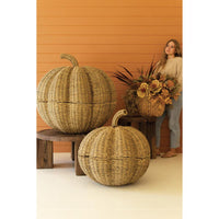 Two large woven pumpkin-shaped decorative containers with lids are displayed on wooden stools in front of an orange wood-paneled wall. A woman stands behind holding a floral arrangement, with one pumpkin container in the foreground and another larger one behind it.