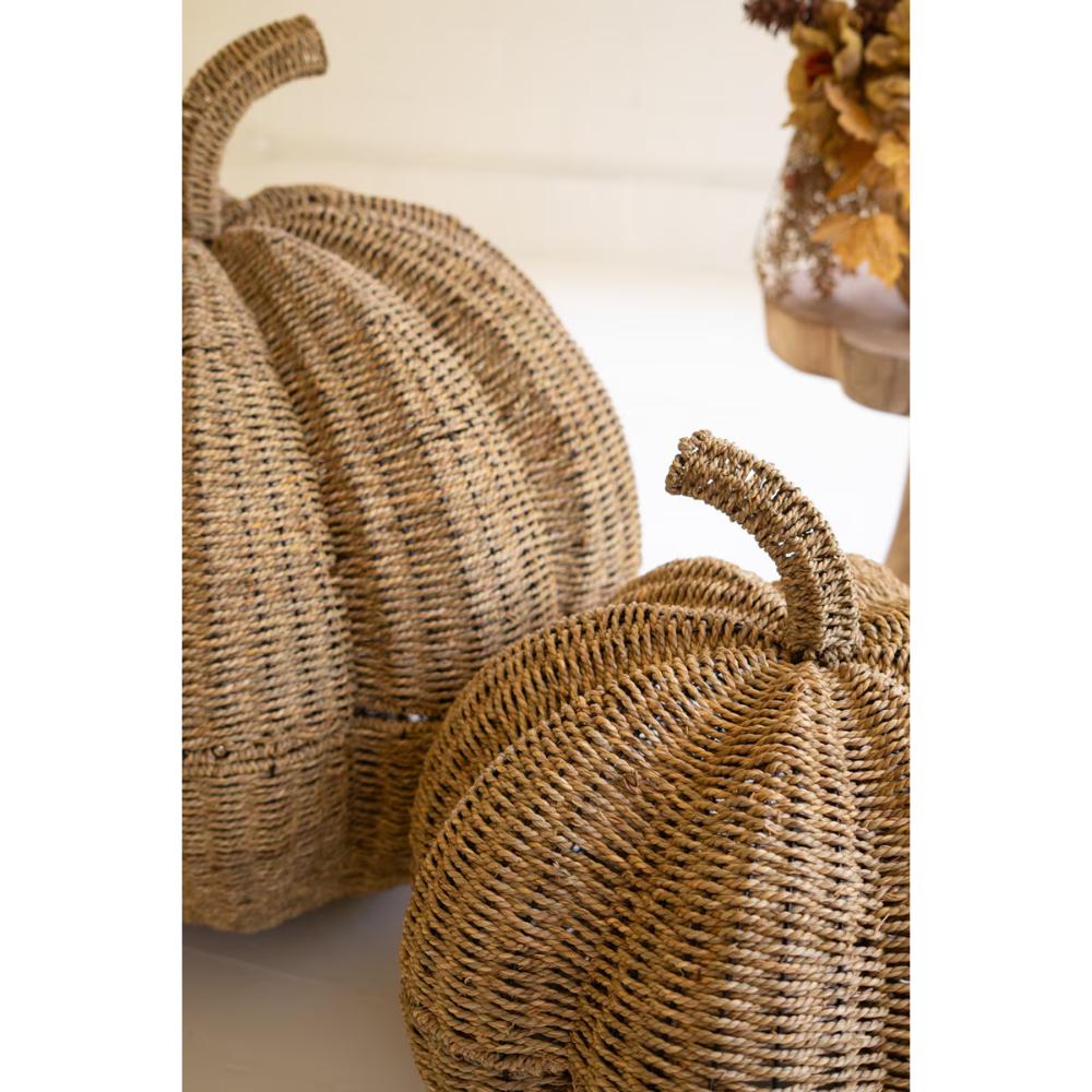 Two woven natural-fiber decorative pumpkins with short stems, shown in a warm neutral indoor setting with a blurred dried floral arrangement in the background.
