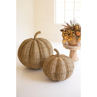 Two large woven seagrass pumpkins in natural tan tones, styled on a white floor next to a small wooden stand holding a dried floral arrangement in a bright room.