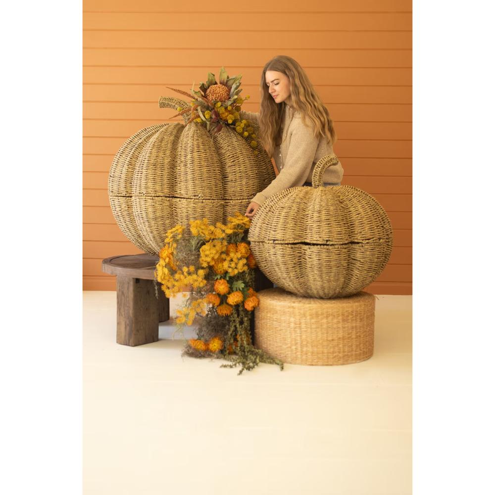 A woman in a beige sweater sits beside two large woven pumpkin-shaped poufs or baskets on a studio set with a warm orange wood-paneled background and autumn floral decor.
