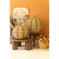 Two large woven seagrass pumpkins with lids, styled on rustic wooden stools against an orange wall with a landscape painting and a vase of autumn flowers.