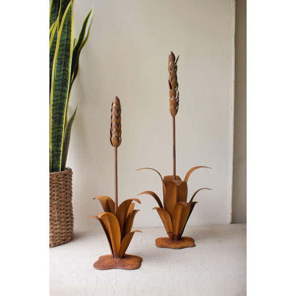 Two decorative metal plant sculptures on a white surface with a plant in the background.