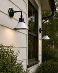 Two Surya Campo Wall Sconces are mounted on a beige house near a window and lush green shrubs.