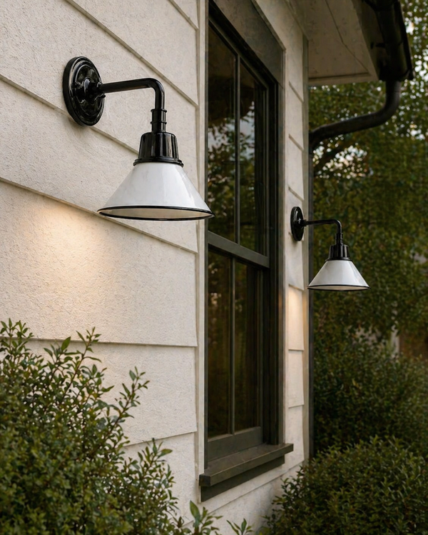 Two Surya Campo Wall Sconces are mounted on a beige house near a window and lush green shrubs.