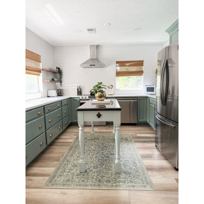 A modern kitchen with green cabinets, stainless appliances, a small island, and a Spicher & Company Joy of Earth vinyl mat.