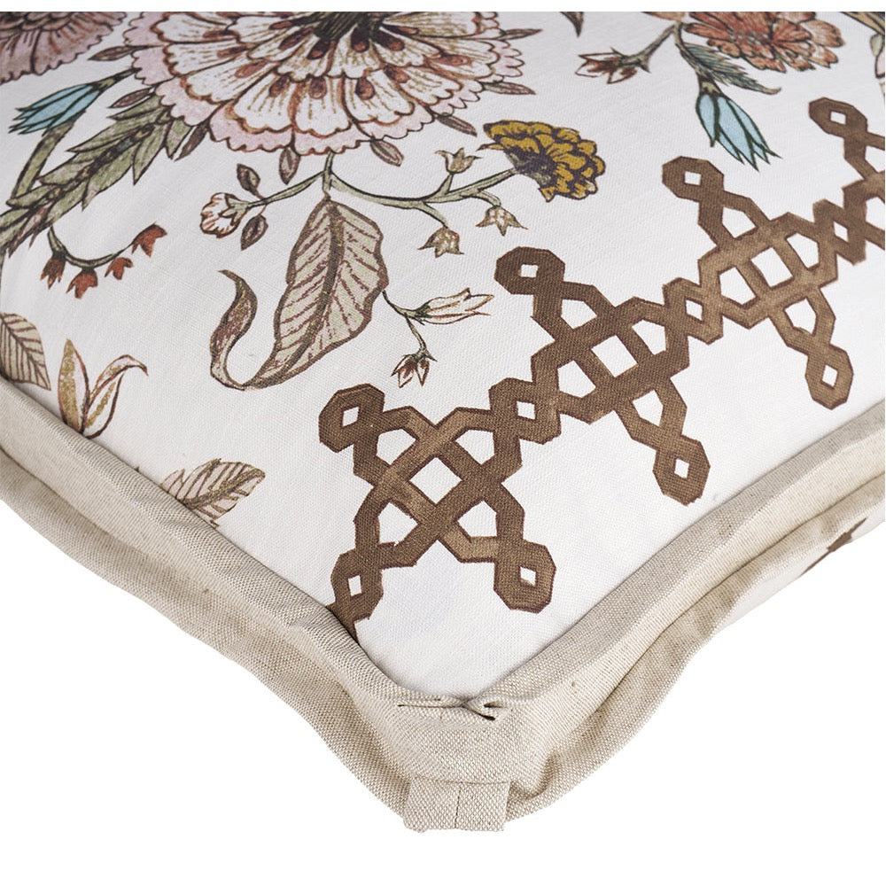 Close-up of a decorative throw pillow corner showing a cream fabric base with a floral print in muted pink, green, and yellow, plus a brown geometric embroidered pattern and a beige piped edge.