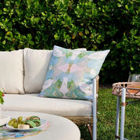 The Laura Park Designs Outdoor Elephant Falls Pillow adds charm to a white outdoor sofa with greenery and a small table.