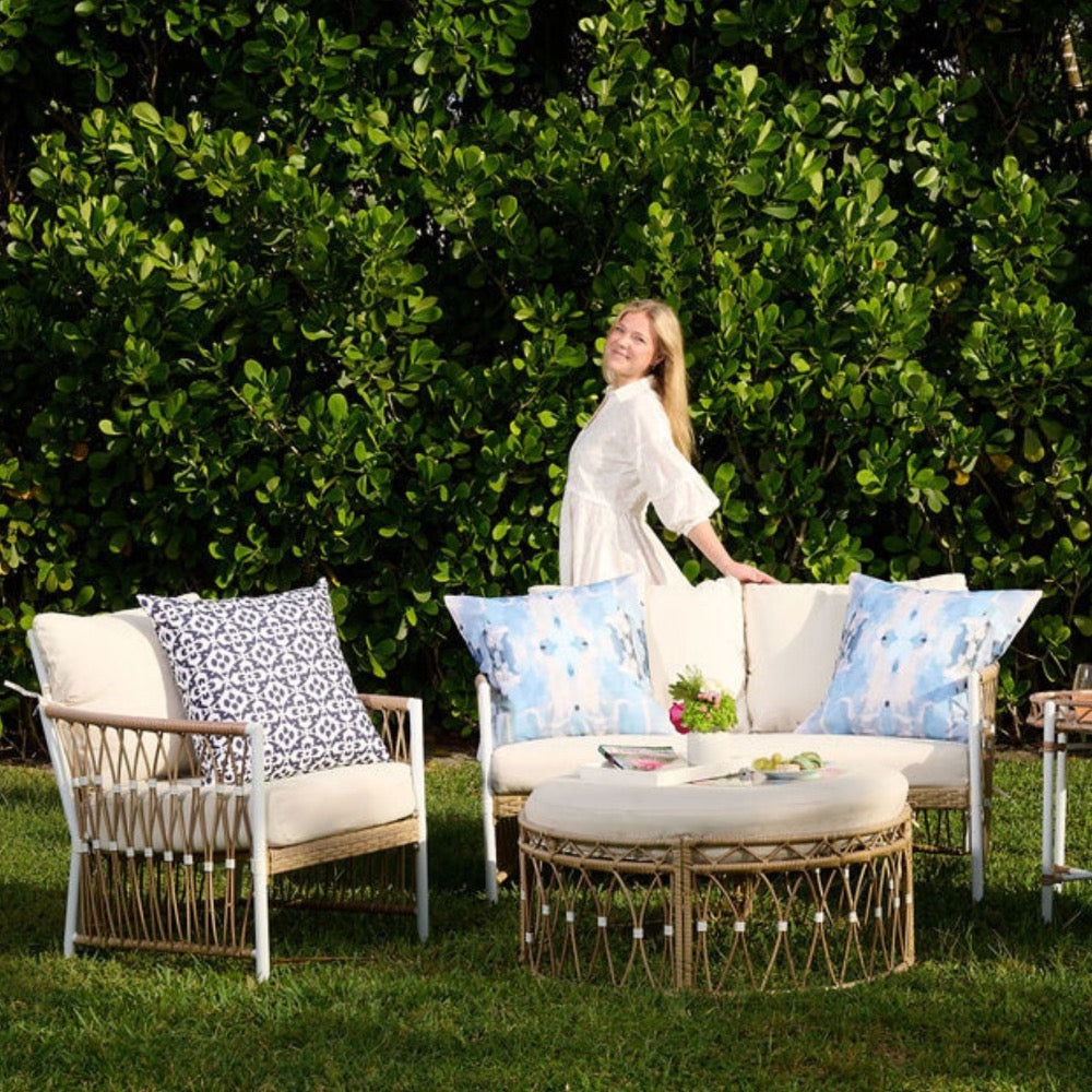 Woman in white dress smiles behind patio set featuring Laura Park Designs' Outdoor Ocean Breeze Pillow.