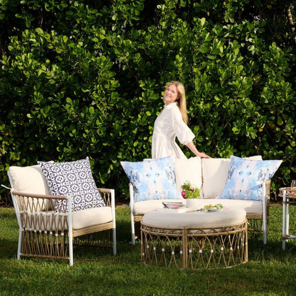 Woman in white dress smiles behind patio set featuring Laura Park Designs' Outdoor Ocean Breeze Pillow.