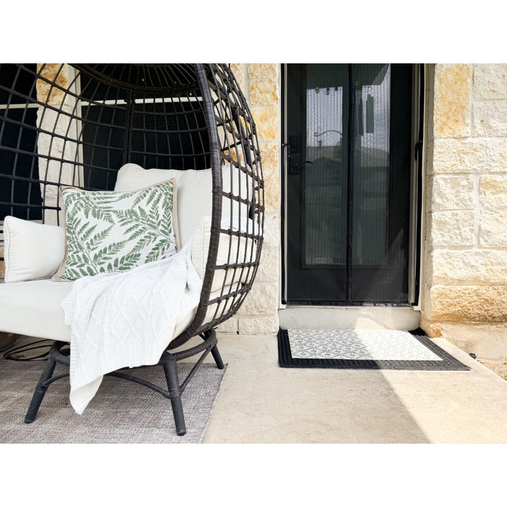 Round wicker chair with white cushions, leafy pillow, throw blanket, and Spicher and Company Clayton Vinyl Floorcloth.