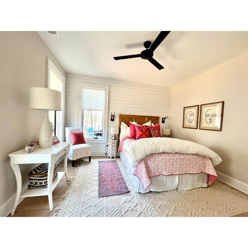 A cozy bedroom featuring a Sanibel Console Table by Phillips Scott, finished in a traditional style with polished brass hardware, complemented by a bed adorned in pink and white bedding and decorative pillows, all under the gentle breeze of a ceiling fan.