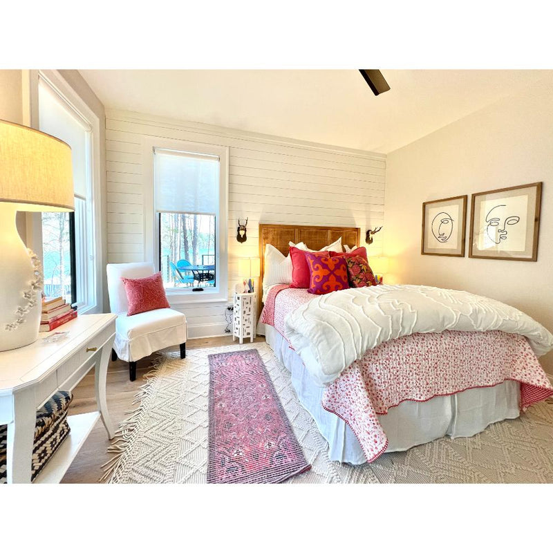 A cozy bedroom features a wooden bed adorned with colorful pillows, two framed artworks, and a window offering views of the trees outside. Adding to the room's charm is the Sanibel Console Table by Phillips Scott, showcasing custom colors and polished brass hardware in a traditional style.