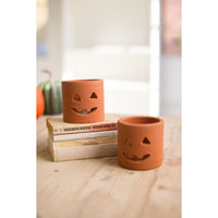 Two terracotta pumpkin-shaped candle holders with carved jack-o'-lantern faces, displayed on a wooden surface with stacked books and a blurred background.