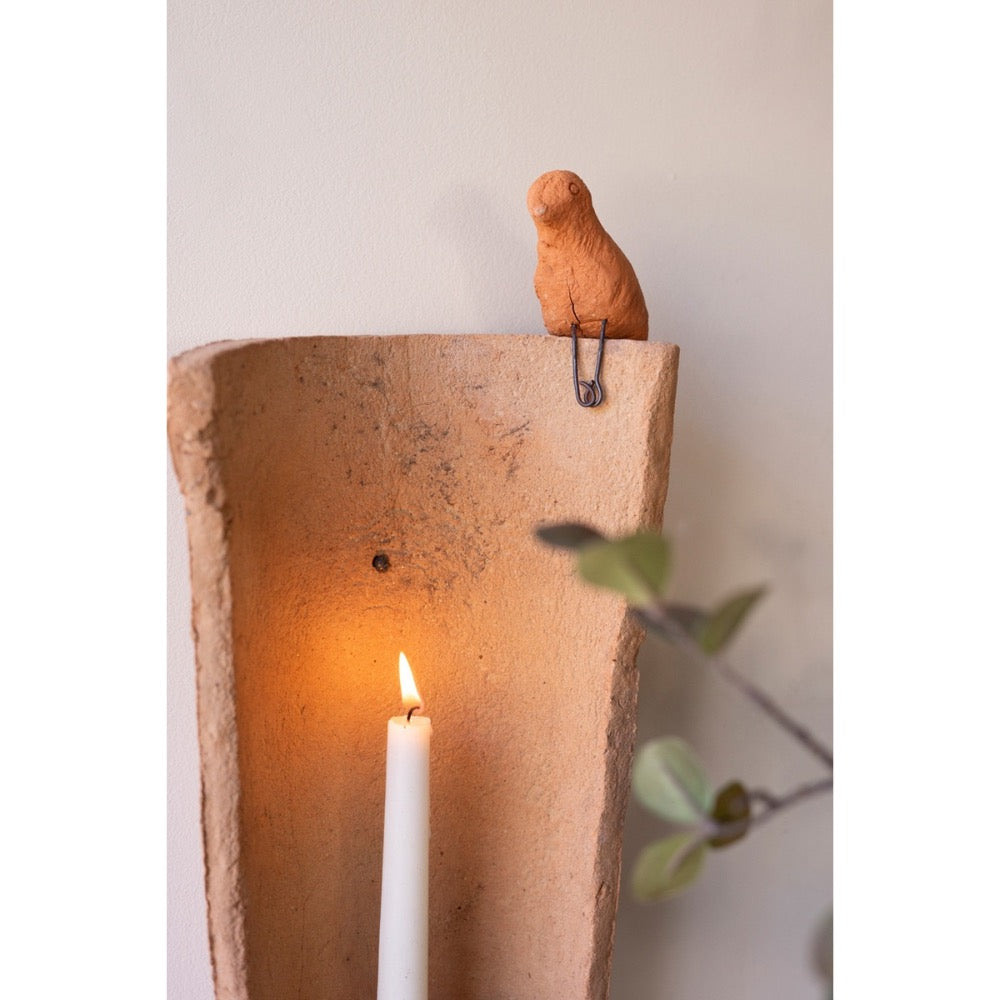 Clay wall sconce with a lit candle inside and a small figurine of a bird on top against a neutral background.