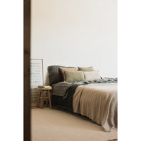 A minimalist bed styled with neutral-toned bedding, including a beige throw blanket, dark gray sheets, and sage green pillows, set in a softly lit bedroom.