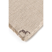 Close-up of a textured beige woven throw corner with fringed edges and a small dark embroidered logo near the tip, shown on a white background.