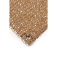 A close-up of a woven throw showing a tan and off-white textured pattern with fringed edges and a small dark embroidered logo near the corner.