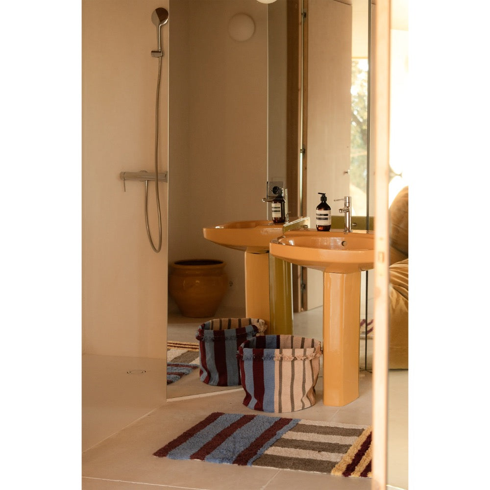 A modern bathroom interior with two light wood pedestal sinks, a wall-mounted shower, striped woven baskets, and a striped bath mat in a warm neutral-toned room.