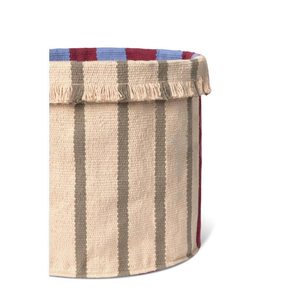 A close-up of a woven storage basket with a beige body, vertical taupe stripes, a fringed top edge, and a striped rim in blue and burgundy on a white background.