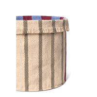A close-up of a woven storage basket with a beige body, vertical taupe stripes, a fringed top edge, and a striped rim in blue and burgundy on a white background.