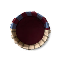 A round woven basket viewed from directly above, featuring a solid deep burgundy center, a blue and burgundy striped outer band, and a beige and brown plaid fringed edge, set against a plain white background.