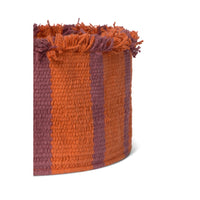 A close-up of a woven basket with vertical stripes in burnt orange and muted mauve, featuring a frayed fringe rim, shown on a white background.