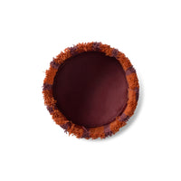 A round decorative throw pillow viewed from directly above, featuring a deep burgundy center and a fringed border in rust orange and dark plum purple.