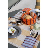 A styled breakfast table setting featuring woven placemats in cream, beige, and rust orange, a striped basket of bread, silver metal dishes, and a small copper cup on a gray table.