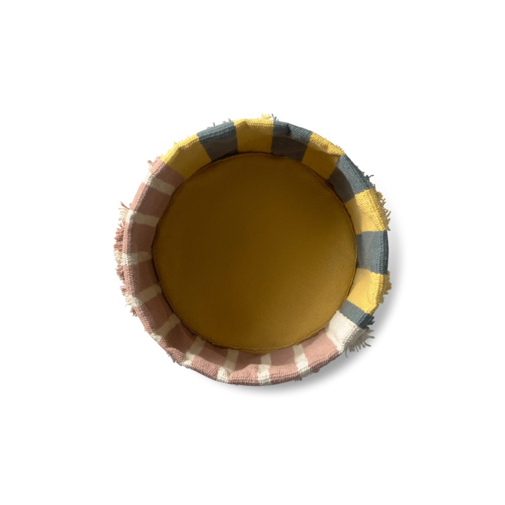 A round woven basket viewed from directly above, featuring a mustard yellow interior and a fringed outer rim with a plaid pattern of yellow, gray, and dusty pink.