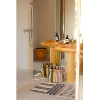 A bathroom scene with two bright yellow pedestal sinks, a large mirror, a striped bath mat, and a yellow ceramic pot in the background.