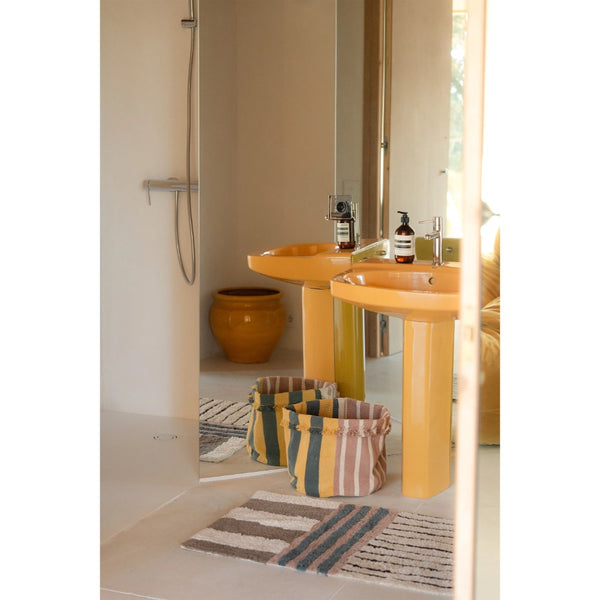 A bathroom scene with two bright yellow pedestal sinks, a large mirror, a striped bath mat, and a yellow ceramic pot in the background.