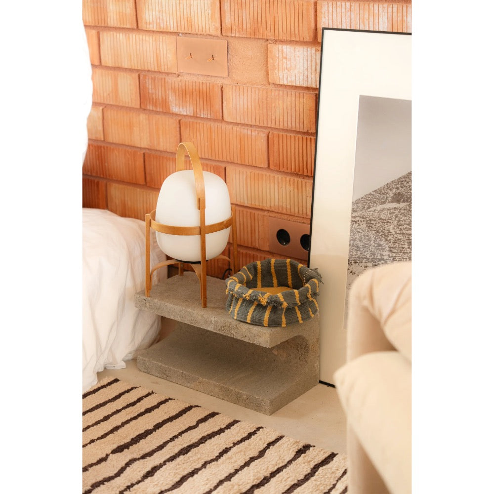 A styled interior scene showing a small concrete side table holding a white spherical lamp in a wooden frame and a striped woven basket, set against a terracotta brick wall with part of a bed and sofa visible.