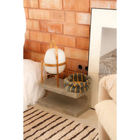 A styled interior scene showing a small concrete side table holding a white spherical lamp in a wooden frame and a striped woven basket, set against a terracotta brick wall with part of a bed and sofa visible.