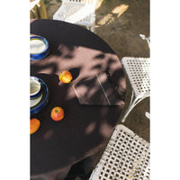 A dark brown round tablecloth draped over an outdoor patio table, featuring dappled shadows from overhead trees. White woven chairs and blue-rimmed ceramic dishes are arranged on the table alongside small oranges.