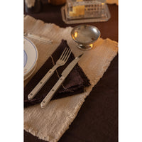 A close-up of silver-toned flatware, including a knife and fork with metal handles, arranged on a dark napkin over a woven beige placemat on a dark table setting.