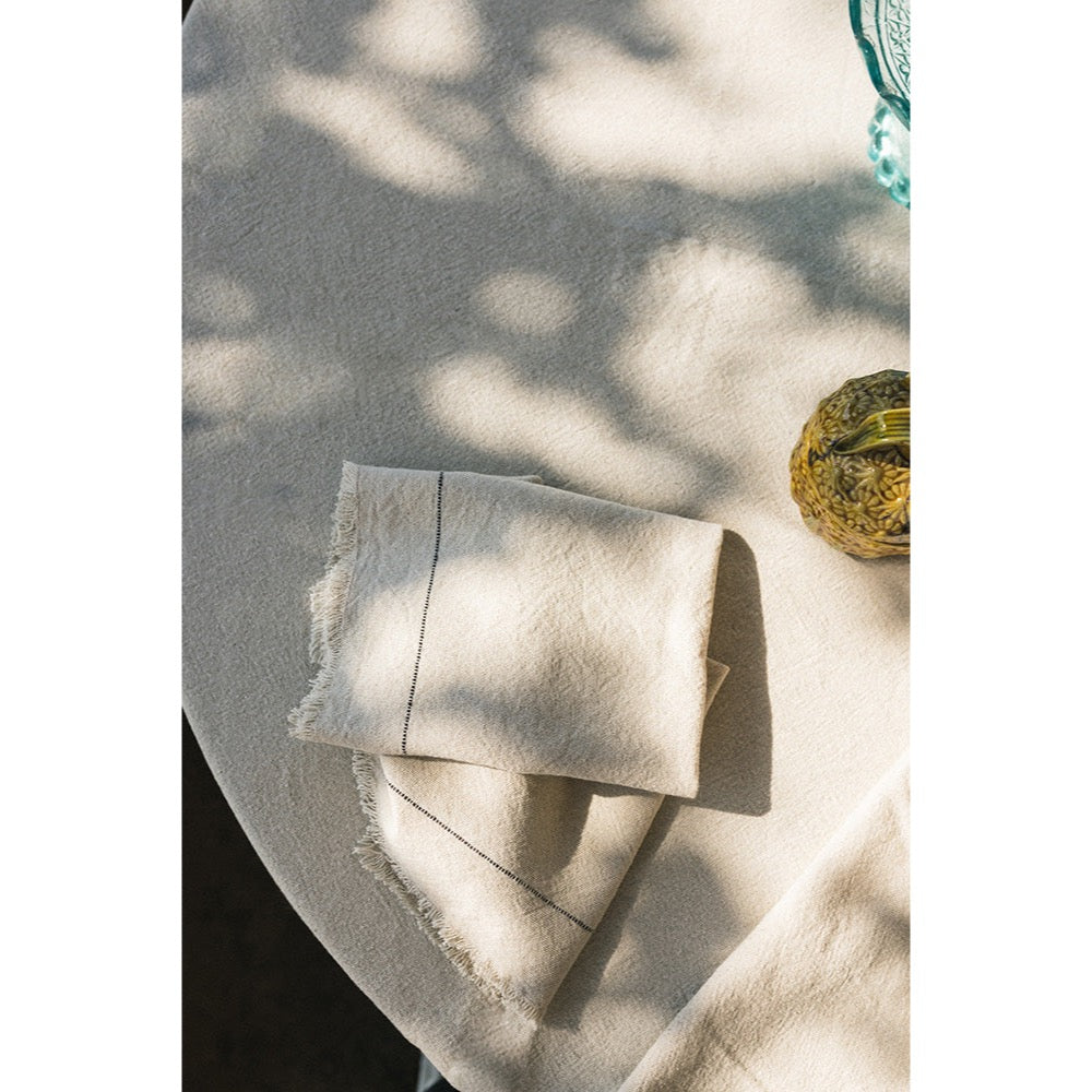 A folded cream-colored linen napkin with subtle stitched details and raw frayed edges, resting on a matching cream linen tablecloth under dappled sunlight.