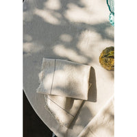 A folded cream-colored linen napkin with subtle stitched details and raw frayed edges, resting on a matching cream linen tablecloth under dappled sunlight.