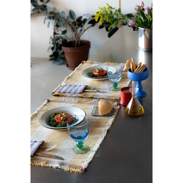 Two woven table runners with fringed edges are styled on a dark tabletop, set with plates, blue and green drinking glasses, breadsticks in a blue holder, and small decorative bottles. Potted plants and flowers sit in the background.