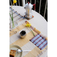 A styled outdoor table setting featuring a woven placemat with a beige and yellow stripe pattern, fringed edges, and a blue-and-red plaid accent patch. A silver metal coffee pot, small metal cup, spoon, and a plate with a slice of cake are arranged on the table, alongside yellow daffodils in a glass vase.