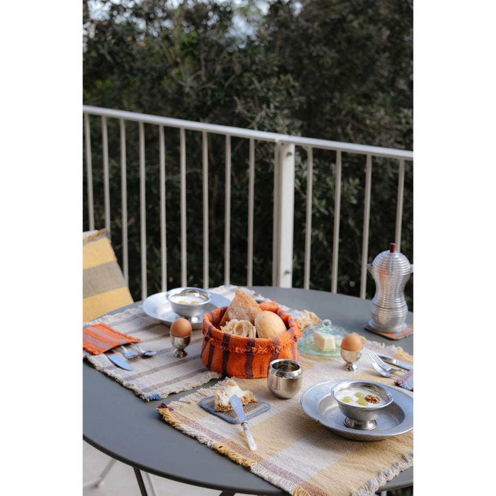 An outdoor dining table set on a balcony with a basket of bread, eggs, plates, cutlery, and a small metal pot, surrounded by green foliage in the background.